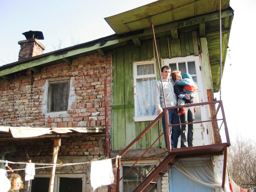 Fotografie cu o familie în fața unei case nefinisate și neizolate, construite improvizat